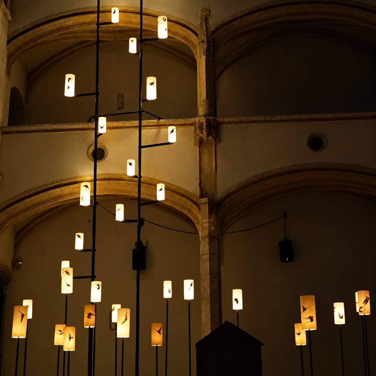 Dans la pénombre d'un édifice ancien aux arcades de pierre, de nombreuses lanternes éclairées décorées de silhouettes d'oiseaux sont fixées sur de hauts mâts.