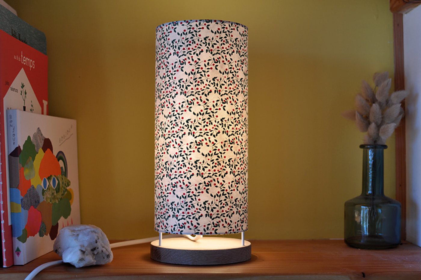 Une lampe cylindrique allumée à l'abat-jour en papier fleuri repose sur une étagère en bois, entre des livres colorés et un vase en verre contenant des fleurs séchées, devant un mur jaune.