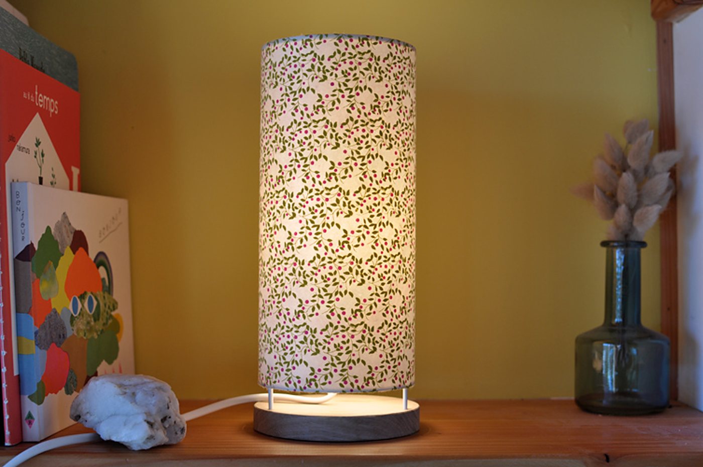 Une lampe de table cylindrique allumée, à abat-jour en papier fleuri et socle en bois, posée sur une étagère près de livres et d'un vase en verre avec des herbes séchées.