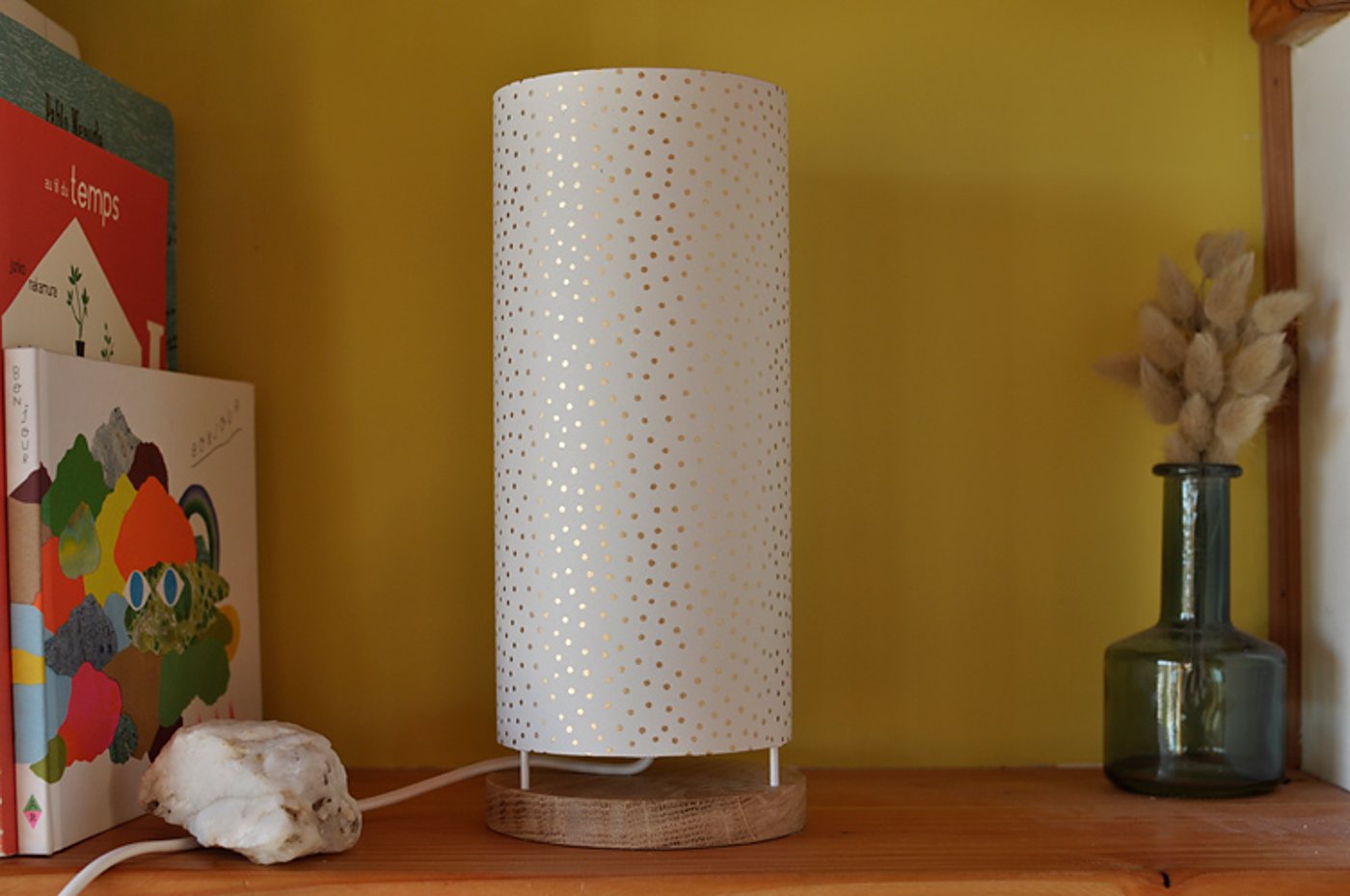 Une lampe de table cylindrique blanche à petits pois dorés, posée sur une base en chêne, sur une étagère en bois devant un mur jaune, entre des livres et un vase en verre avec des herbes séchées.