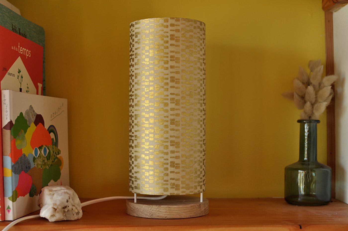 Une lampe cylindrique à abat-jour doré motifié et base en bois clair est posée sur une étagère, entre des livres d'enfants et un vase en verre avec des fleurs séchées.