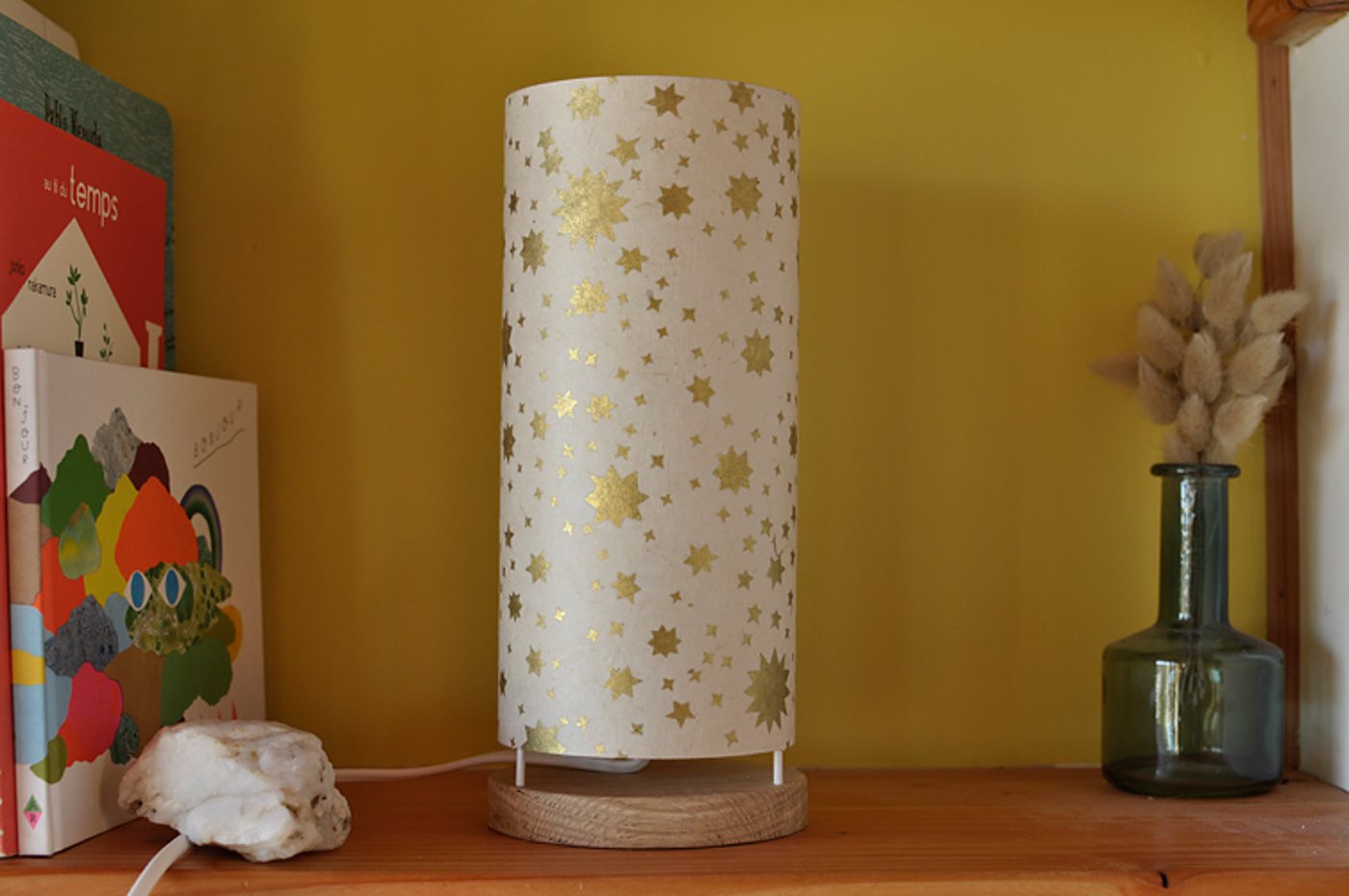 Une lampe cylindrique en papier blanc décoré d'étoiles dorées, sur un socle en bois, est posée sur une étagère près de livres et d'un vase en verre avec des herbes séchées.