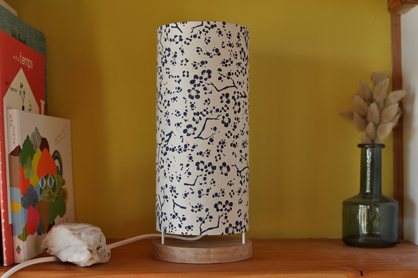 Une lampe cylindrique en papier blanc à motif de fleurs de cerisier bleu marine, posée sur un socle en bois, sur une étagère près de livres et d'un vase en verre avec des fleurs séchées.