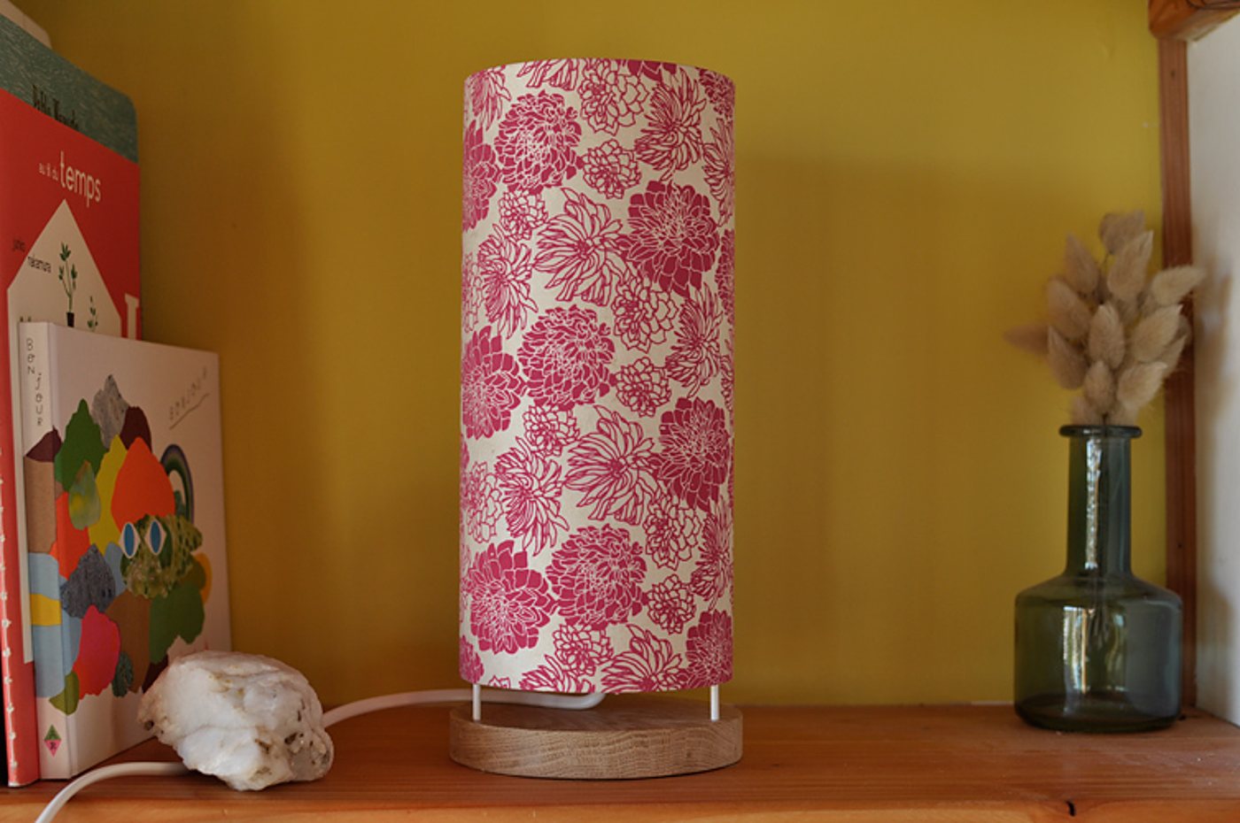 Une lampe cylindrique à abat-jour fleuri rose et blanc posée sur un socle en bois, sur une étagère devant un mur jaune, entre des livres et un vase en verre.