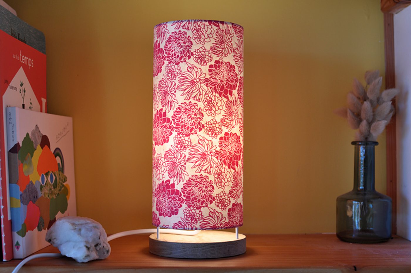 Une lampe cylindrique allumée, au abat-jour blanc orné de fleurs roses, posée sur une étagère en bois entre des livres et un vase en verre avec des herbes séchées.