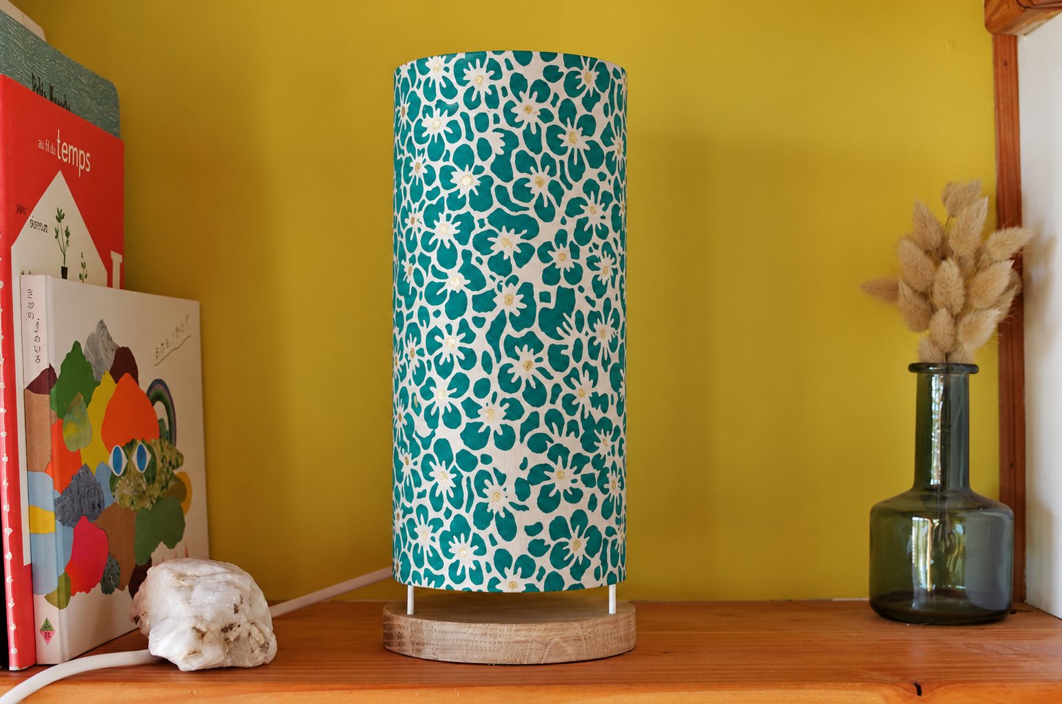 Une lampe cylindrique en tissu à fleurs turquoise et blanches posée sur une étagère en bois, entre des livres et un vase en verre avec des herbes séchées, devant un mur jaune.