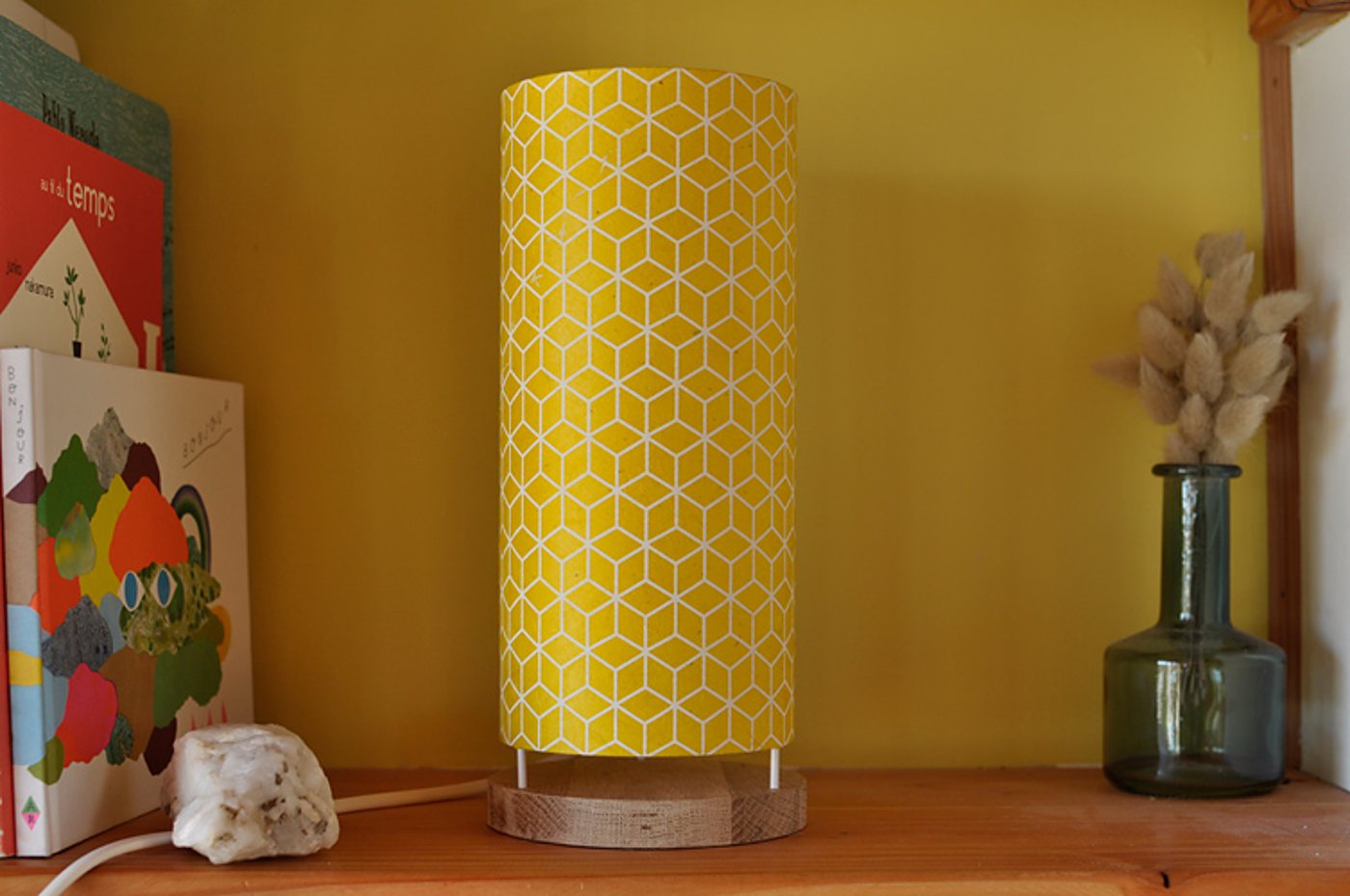 Une lampe cylindrique jaune à motif de cubes blancs, posée sur un socle en bois, entre des livres illustrés et un vase vert avec des herbes séchées.