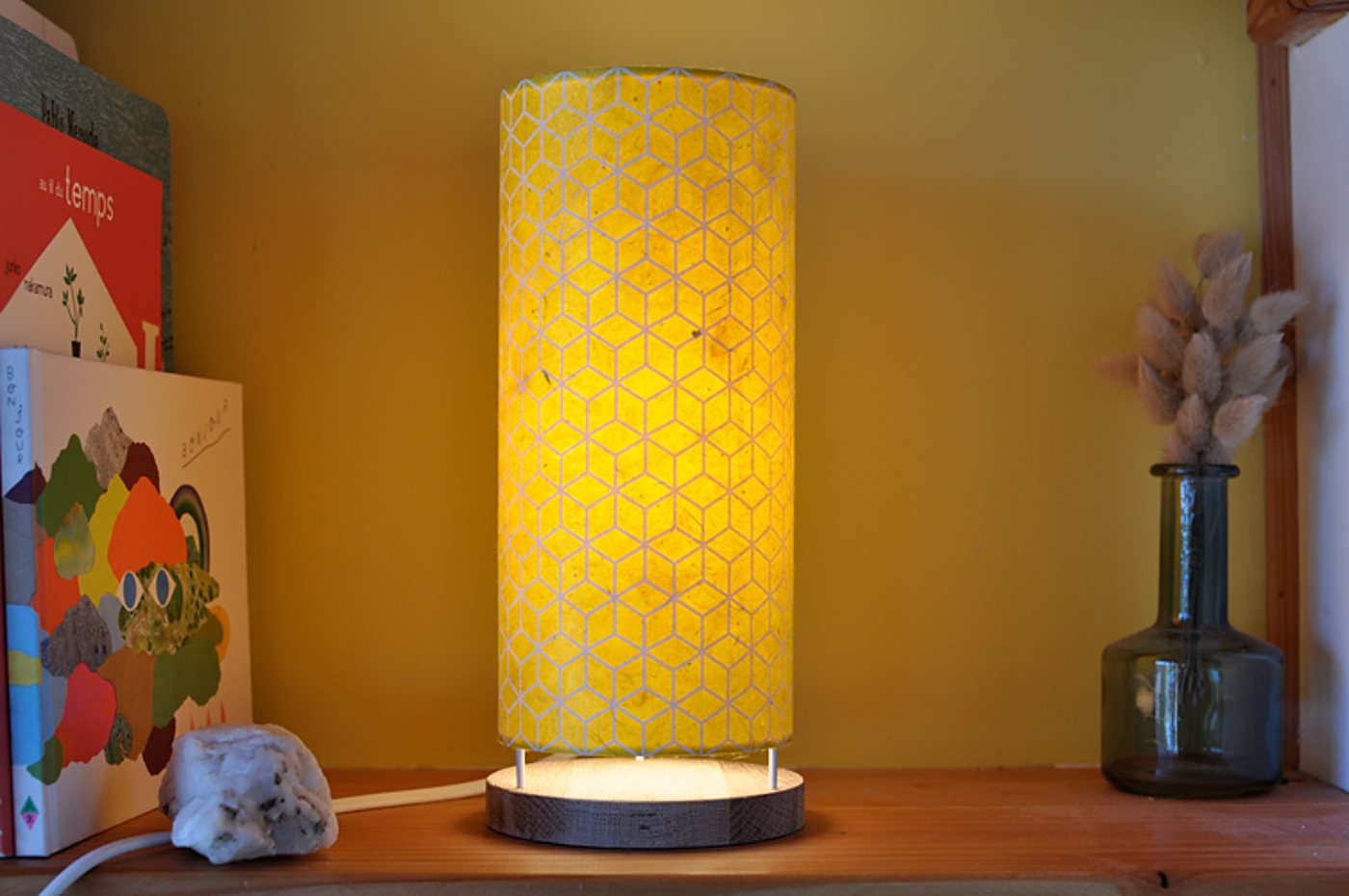 Une lampe cylindrique jaune à motif de cubes géométriques, allumée sur une étagère en bois, entre des livres et un vase en verre contenant des fleurs séchées.