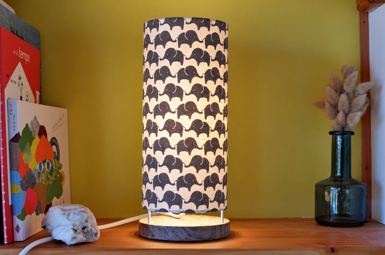 Une lampe cylindrique allumée au abat-jour blanc orné d'éléphants gris, posée sur une étagère en bois près de livres et d'un vase en verre avec des fleurs séchées.