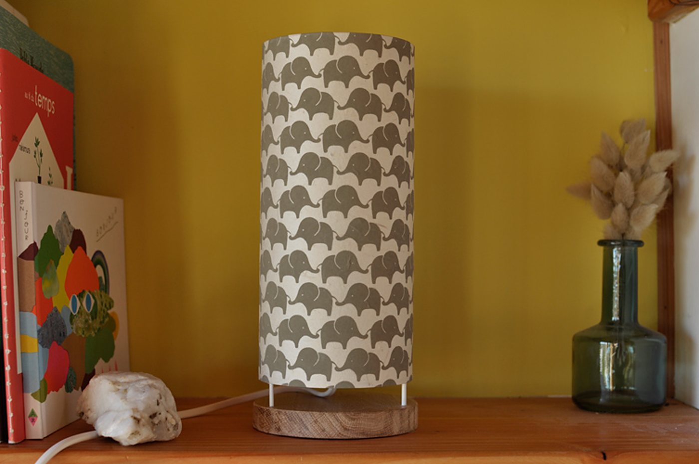 Une lampe cylindrique à motif d'éléphants gris sur socle en bois est posée sur une étagère, entre des livres d'enfants et un vase en verre avec des fleurs séchées.