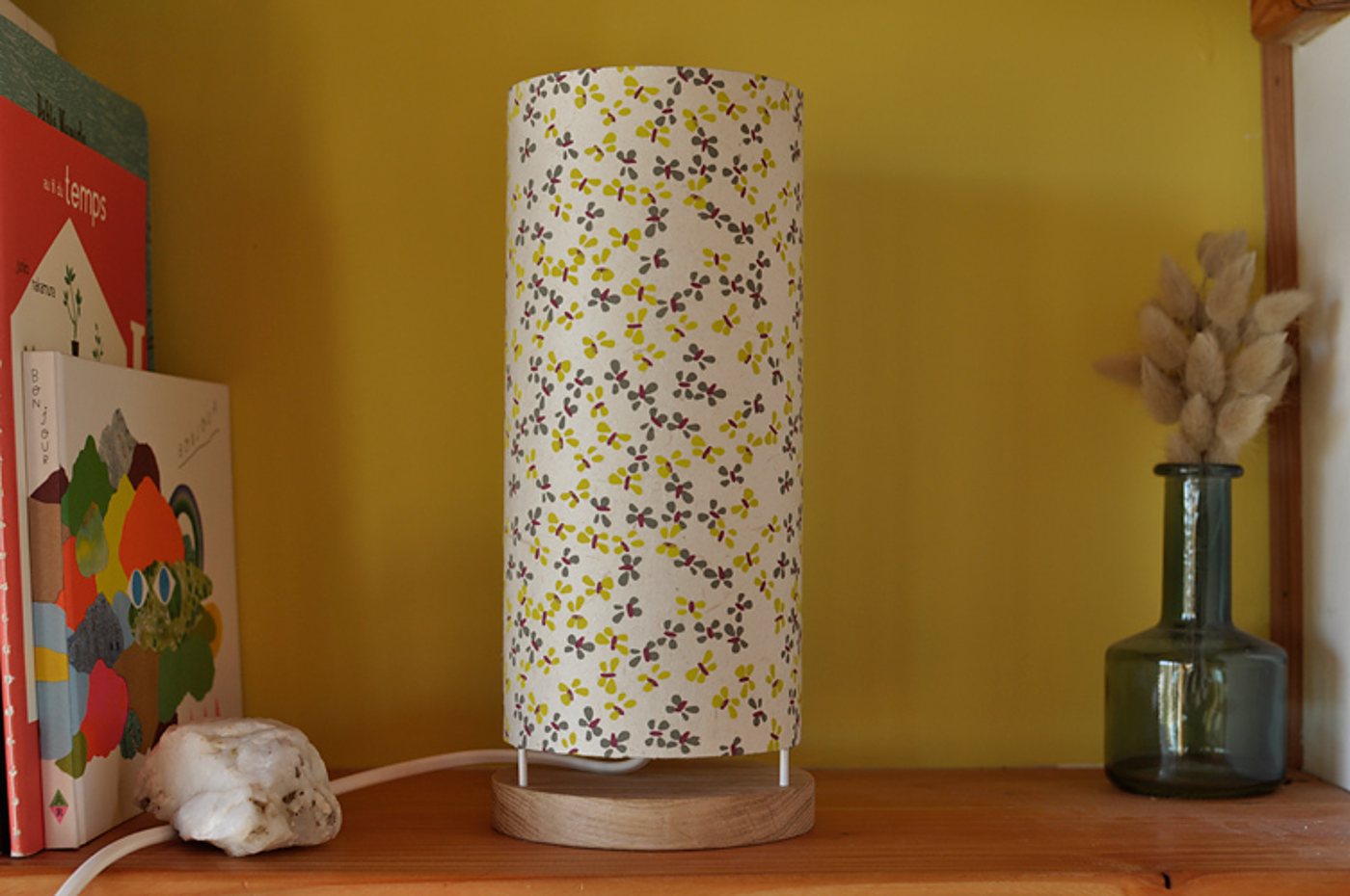 Une lampe de chevet cylindrique à abat-jour fleuri jaune et gris, posée sur une base en bois, sur une étagère devant un mur jaune, entre des livres et un vase en verre avec des fleurs séchées.