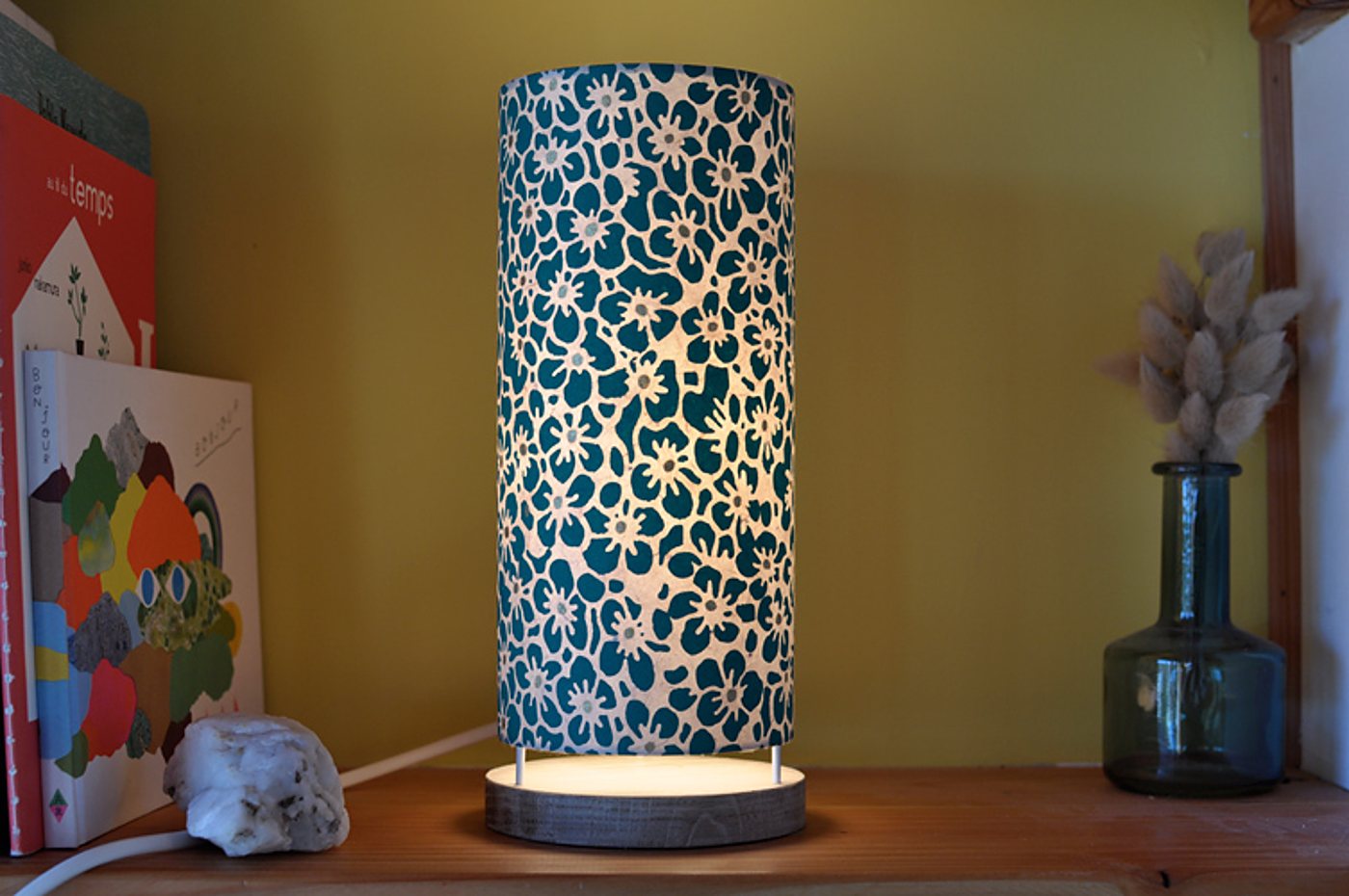 Une lampe cylindrique allumée au abat-jour à fleurs bleu canard et blanches, posée sur une étagère en bois près de livres et d'un vase en verre avec des plantes séchées.