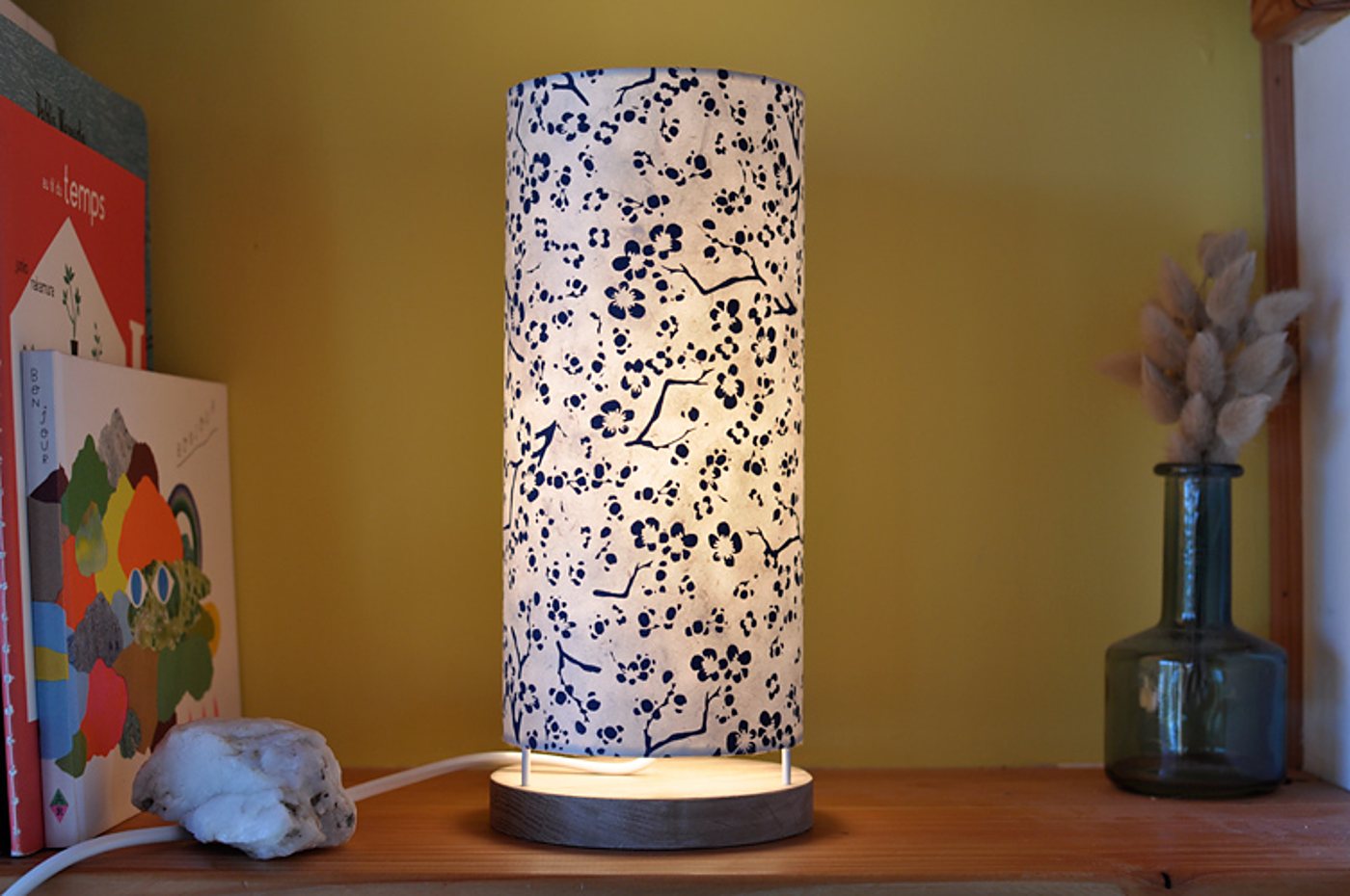 Une lampe cylindrique allumée, à abat-jour blanc orné de fleurs bleu marine, posée sur une étagère en bois près de livres et d'un vase en verre avec des herbes séchées.
