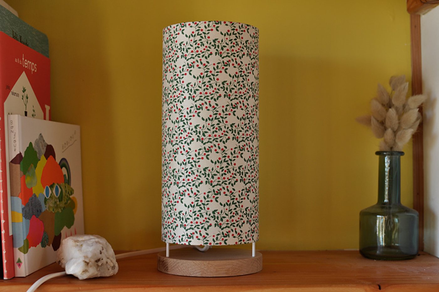 Une lampe cylindrique à abat-jour fleuri et socle en bois est posée sur une étagère, entre des livres illustrés et un vase en verre avec des herbes séchées, devant un mur jaune.