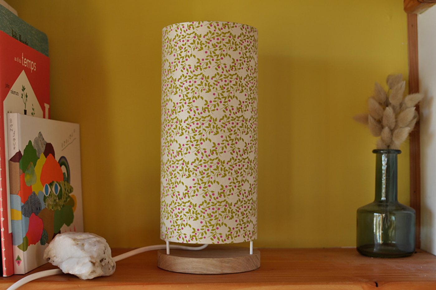 Une lampe cylindrique à abat-jour fleuri vert et rose, posée sur une base en bois sur une étagère, entre des livres et un vase en verre avec des herbes séchées.