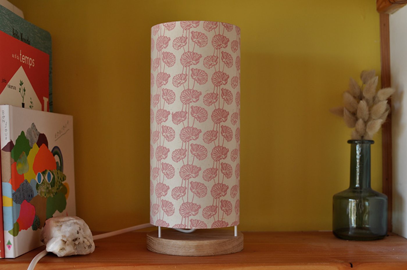 Une lampe cylindrique au abat-jour crème à motifs roses, posée sur un socle en bois, sur une étagère jaune entre des livres et un vase en verre avec des fleurs séchées.