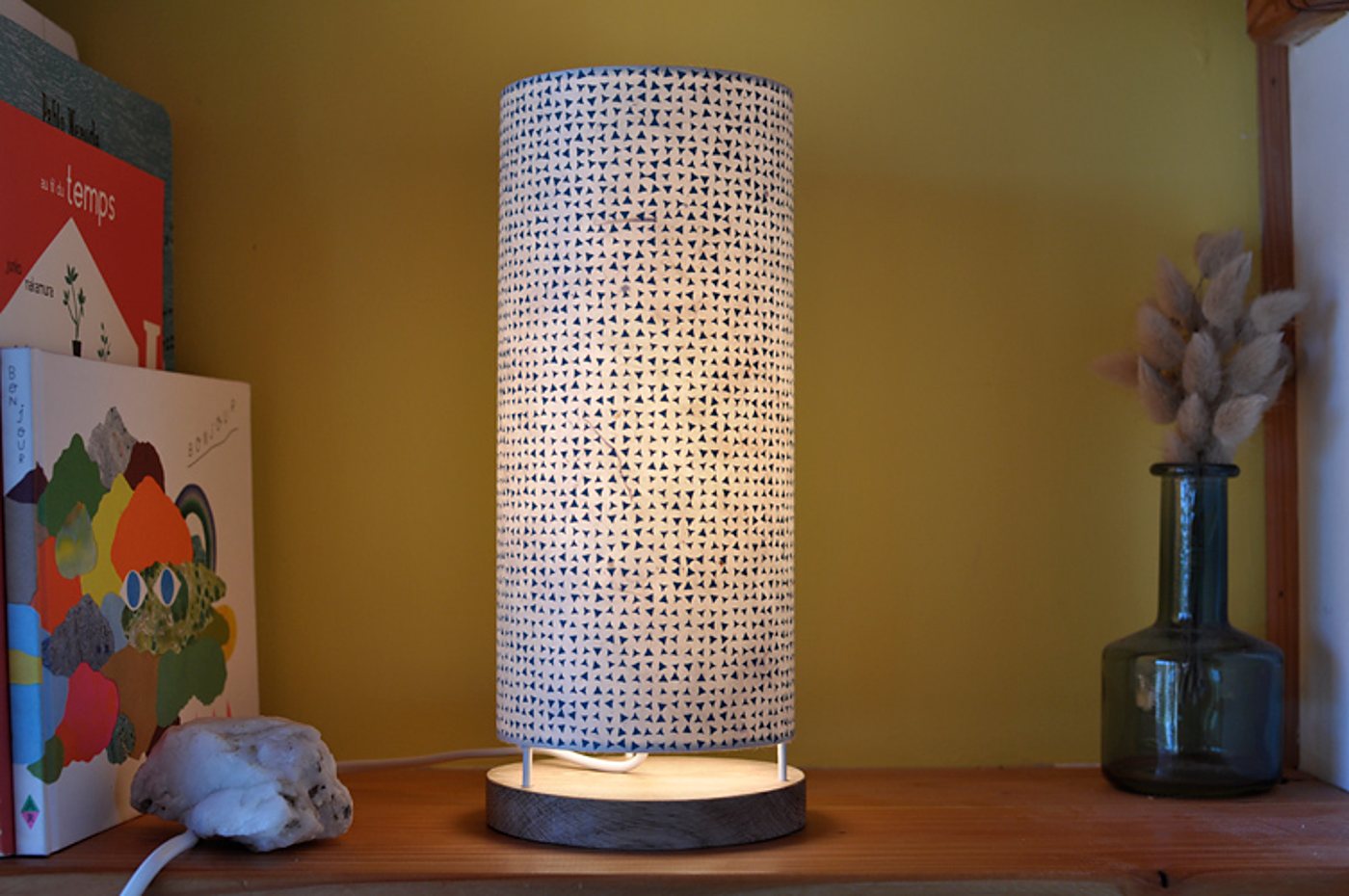 Une lampe cylindrique allumée à abat-jour blanc orné de petits triangles bleus, posée sur une étagère en bois près de livres et d'un vase en verre avec des fleurs séchées.