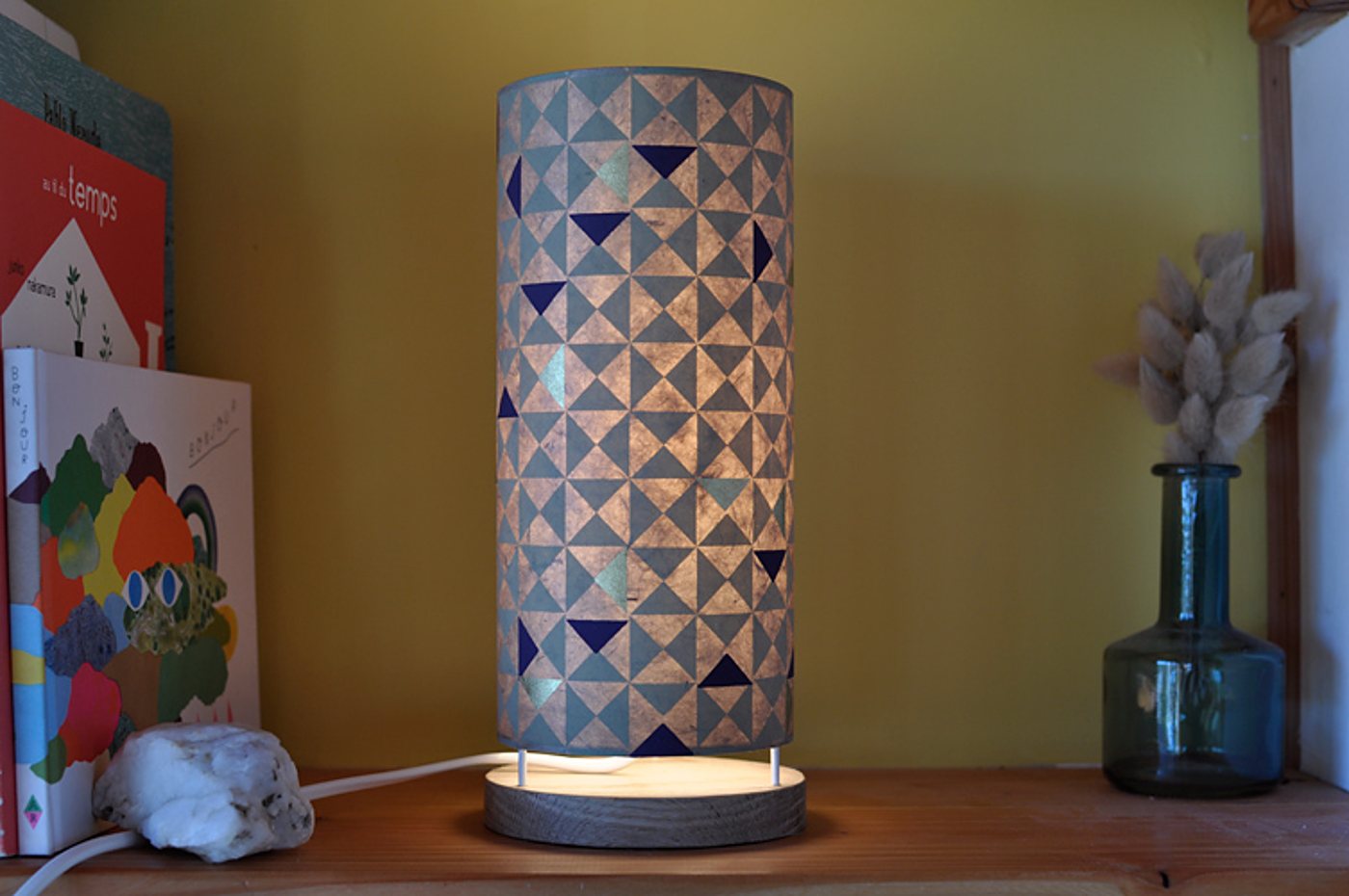 Une lampe cylindrique allumée au abat-jour à motifs de triangles bleus, posée sur une étagère en bois près de livres et d'un vase en verre bleu avec des fleurs séchées.