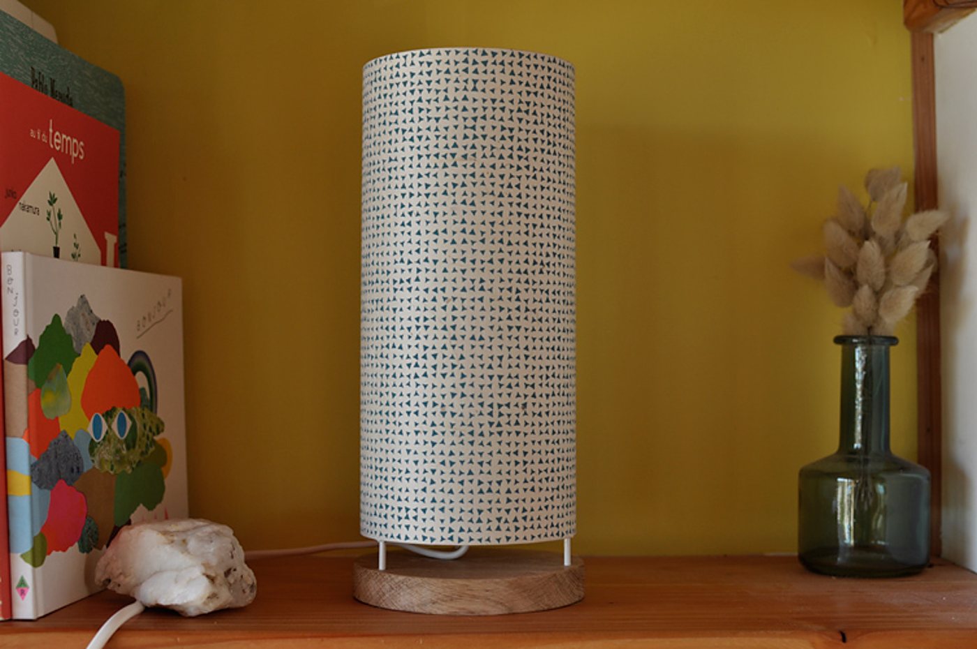 Une lampe cylindrique à abat-jour motifs triangles bleus sur socle en bois, posée sur une étagère entre des livres et un vase en verre avec des fleurs séchées.