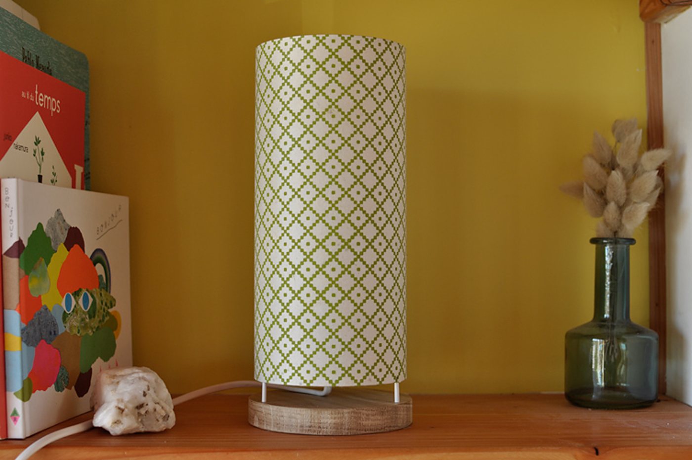 Une lampe cylindrique à motif losanges verts sur socle en bois est posée sur une étagère, entre des livres et un vase en verre avec des herbes séchées.
