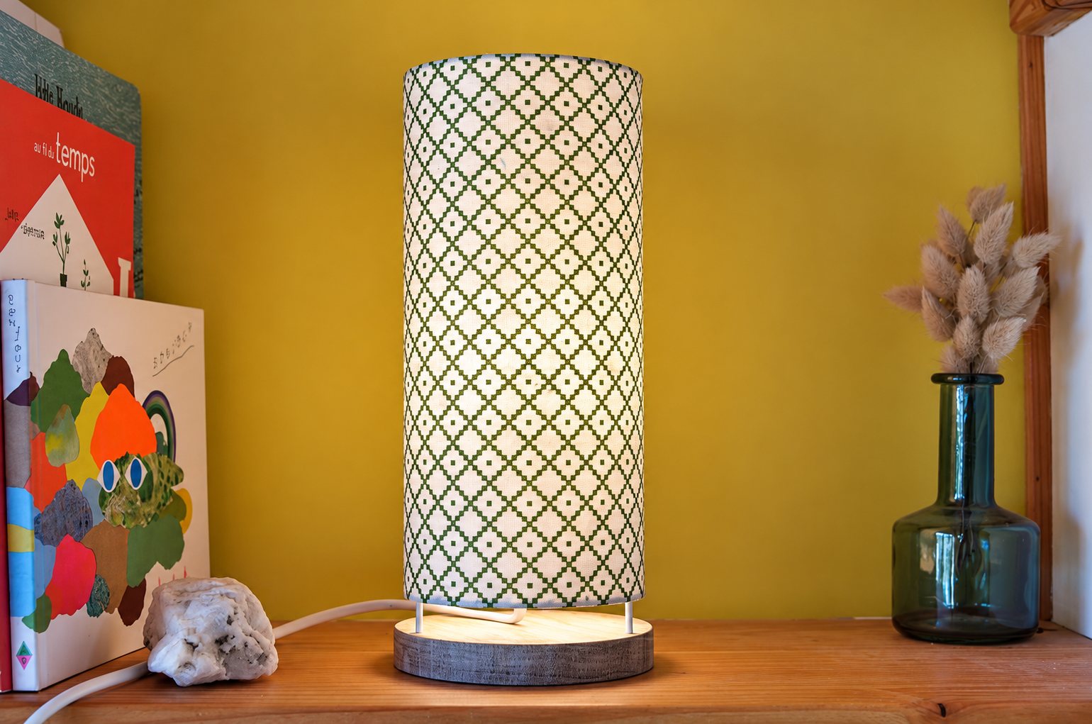 Une lampe de chevet cylindrique allumée, au abat-jour blanc à motif losangé vert, posée sur une étagère en bois entre des livres et un vase en verre avec des fleurs séchées.