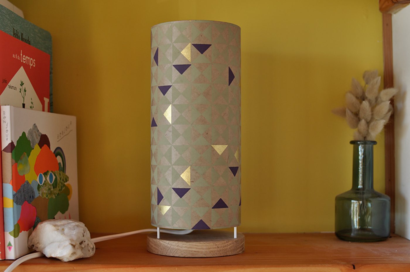 Une lampe de table cylindrique à abat-jour géométrique vert, violet et doré, posée sur une étagère en bois près de livres et d'un vase en verre avec des fleurs séchées.