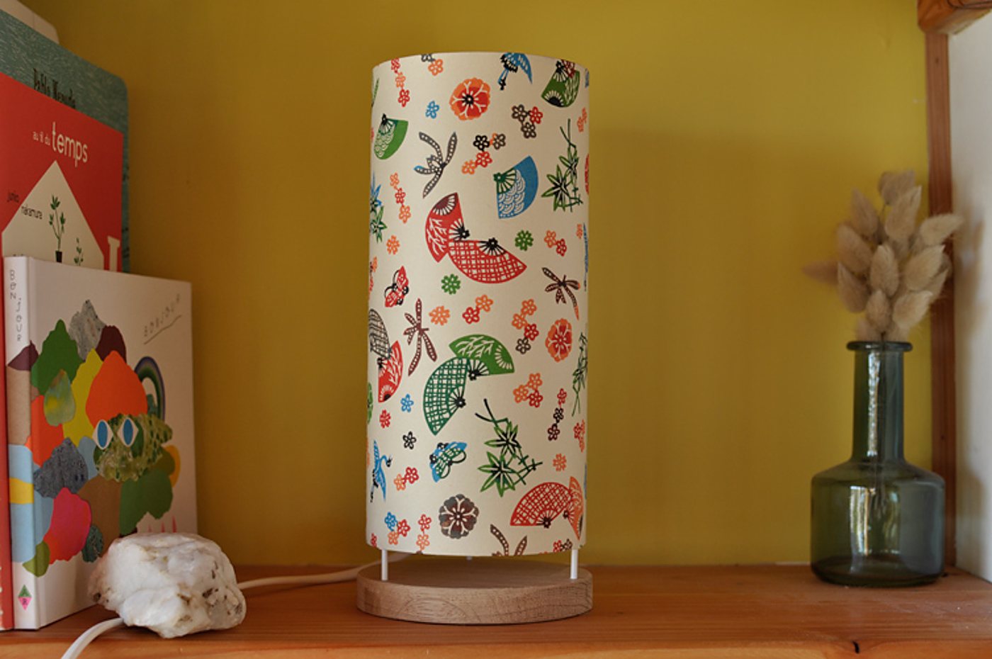 Une lampe de chevet cylindrique au abat-jour fleuri de motifs d'éventails japonais est posée sur une étagère en bois devant un mur jaune, à côté de livres et d'un vase en verre avec des fleurs séchées.
