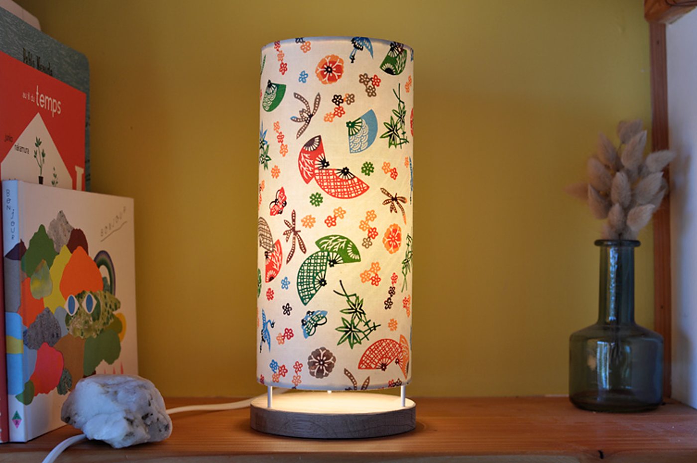 Une lampe cylindrique allumée, au abat-jour orné d'éventails et de fleurs colorés, est posée sur une étagère en bois près de livres et d'un vase.