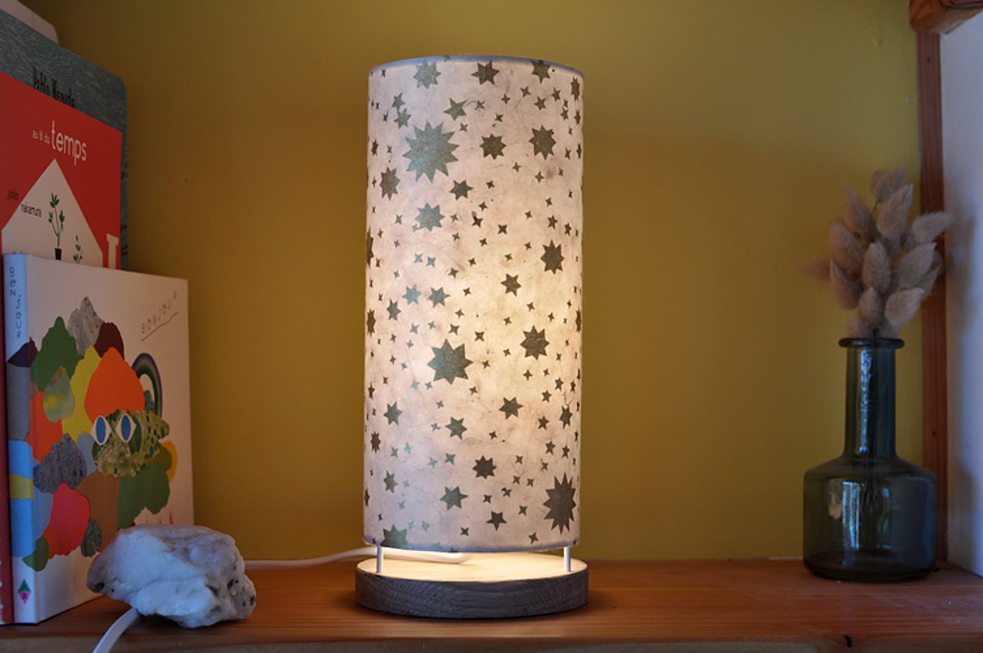 Une lampe cylindrique allumée en papier clair décoré d'étoiles vertes, posée sur une étagère en bois à côté de livres et d'un vase en verre avec des fleurs séchées.
