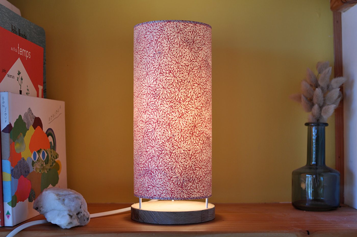Une lampe cylindrique allumée, à abat-jour en papier rose à motifs de fleurs, posée sur une étagère en bois près de livres et d'un vase en verre.