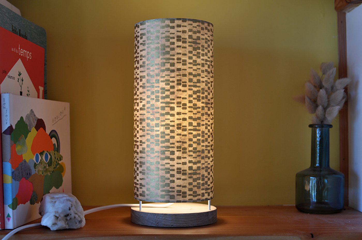 Une lampe de table cylindrique allumée, à l'abat-jour en papier à motifs gris et à base en bois, posée sur une étagère entre des livres et un vase en verre bleu avec des fleurs séchées.