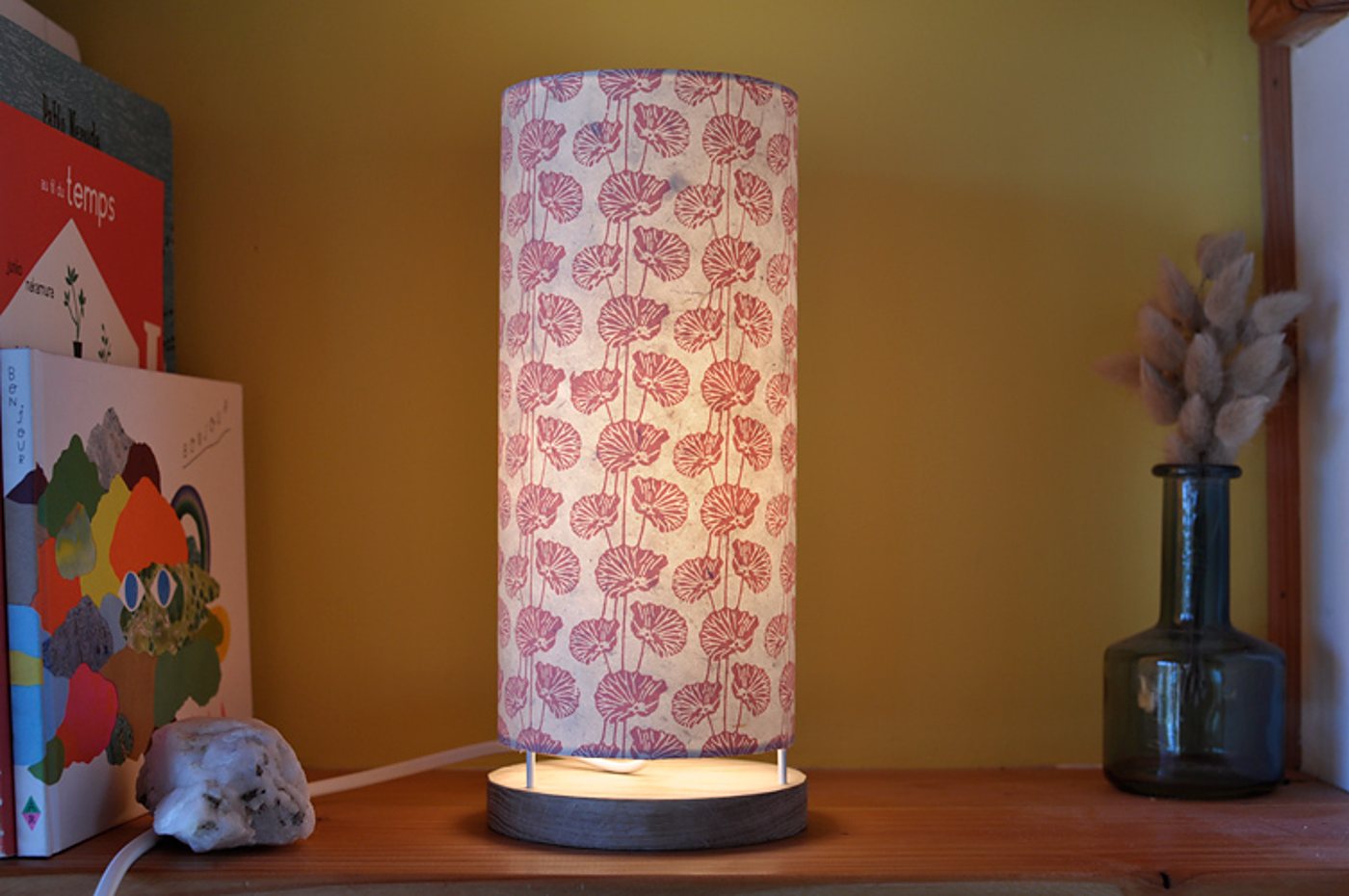 Une lampe de table cylindrique allumée, en papier à motifs floraux roses, posée sur une étagère en bois à côté de livres et d'un vase en verre avec des fleurs séchées.