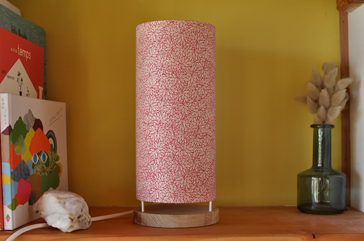 Une lampe de table cylindrique en papier rose à motifs de feuilles blanches, posée sur une base en bois, sur une étagère près de livres pour enfants et d'un vase en verre avec des fleurs séchées.
