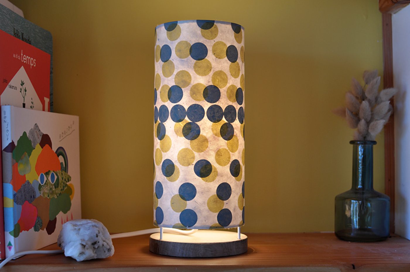 Une lampe de table cylindrique allumée, ornée de pois bleus et jaunes, posée sur une étagère en bois à côté de livres et d'un vase en verre avec des fleurs séchées.