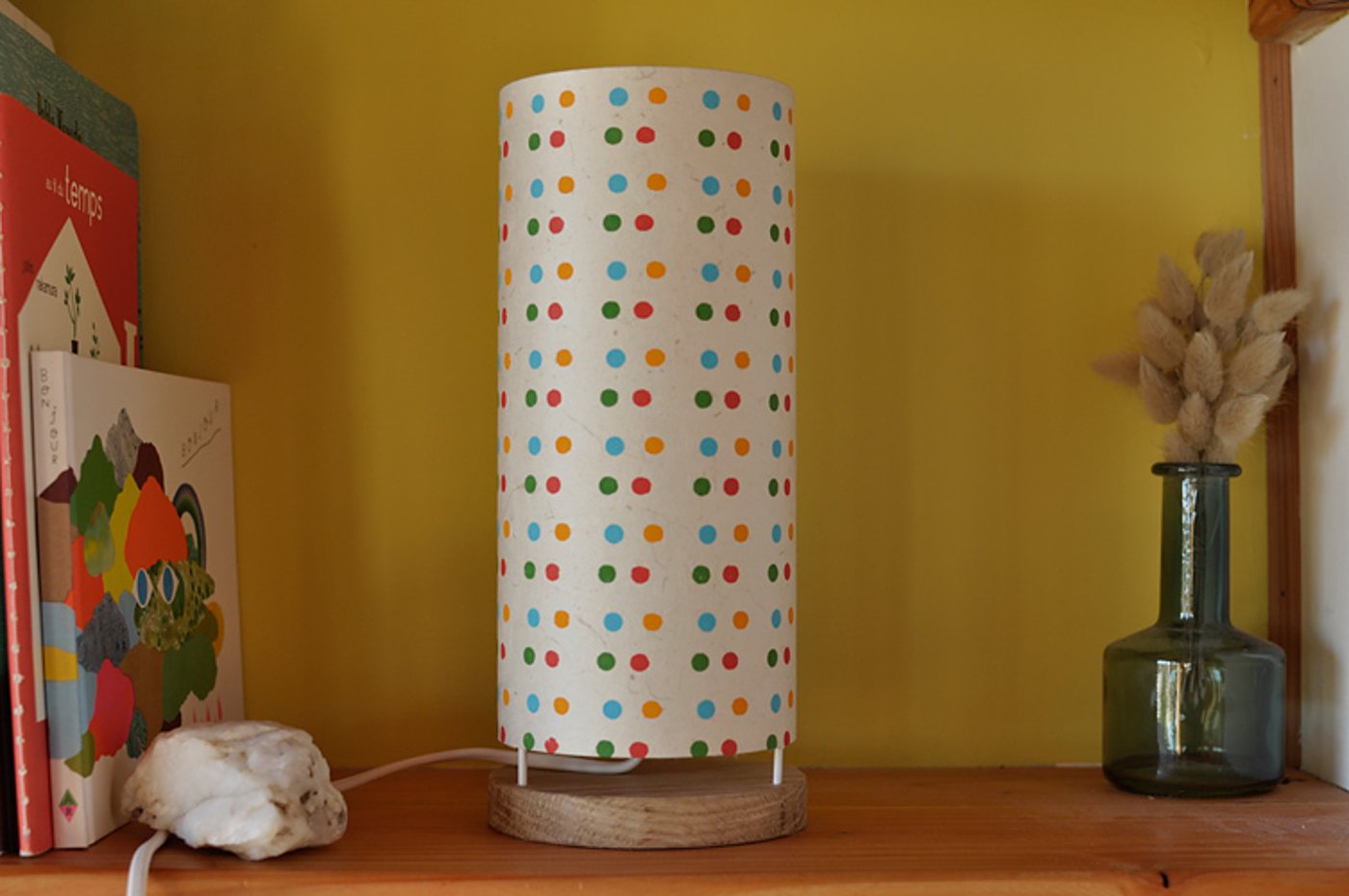 Une lampe cylindrique en papier à pois multicolores sur socle en bois, posée sur une étagère devant un mur jaune, entre des livres et un vase en verre avec des herbes séchées.