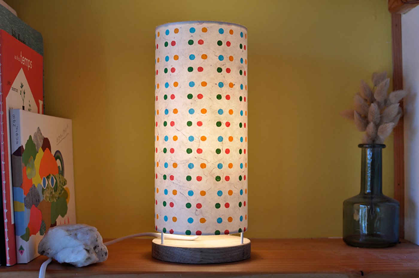 Une lampe cylindrique allumée, à abat-jour en papier blanc orné de pois colorés et socle en bois, est posée sur une étagère près de livres et d'un vase en verre avec des herbes séchées.