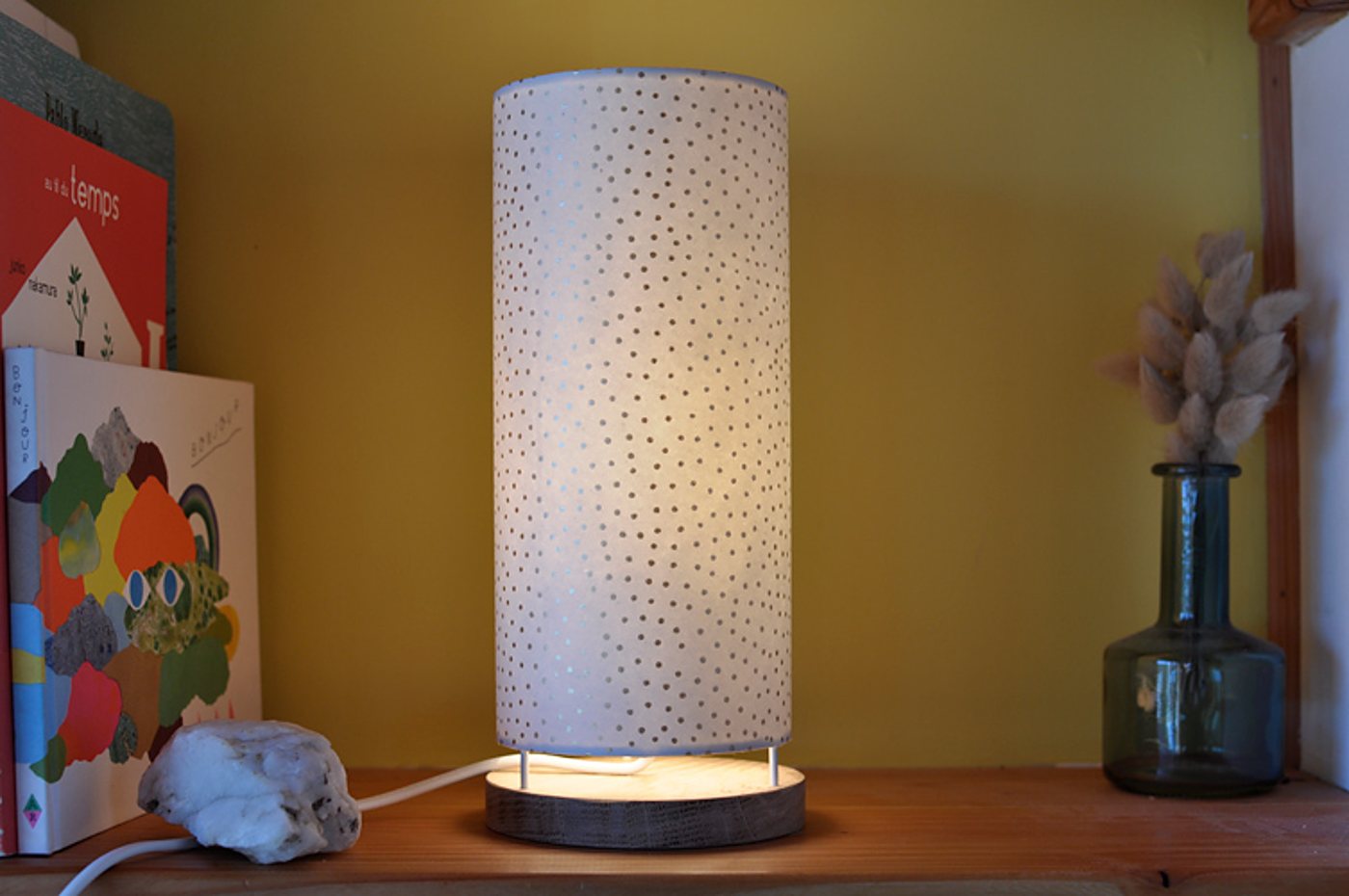 Une lampe de table cylindrique à abat-jour blanc moucheté de pois, allumée sur une étagère en bois, entre des livres et un vase en verre avec des fleurs séchées.