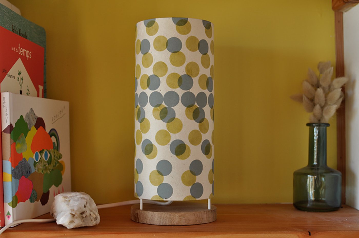 Une lampe cylindrique en papier à pois gris et jaunes, posée sur une base en bois, est placée sur une étagère entre des livres illustrés et un vase en verre avec des herbes séchées.
