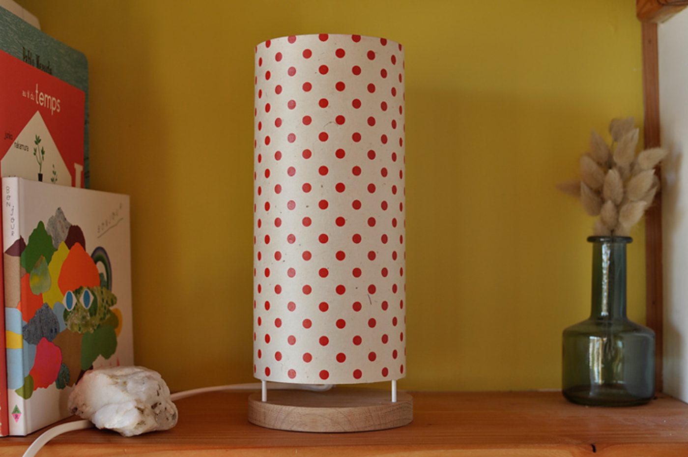 Une lampe cylindrique en papier blanc à pois rouges, sur un socle en bois clair, est posée sur une étagère devant un mur jaune, entre des livres d'enfants et un vase en verre avec des fleurs séchées.