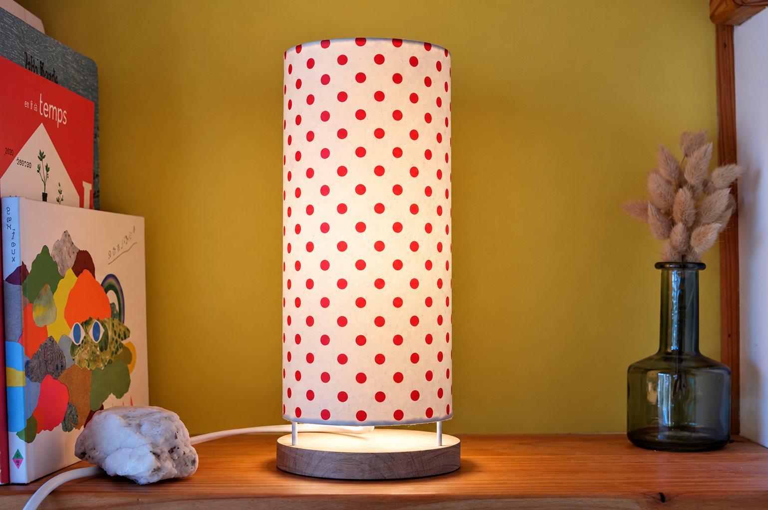 Une lampe cylindrique en papier blanc à pois rouges, allumée sur une étagère en bois, entre des livres colorés et un vase en verre avec des herbes séchées.