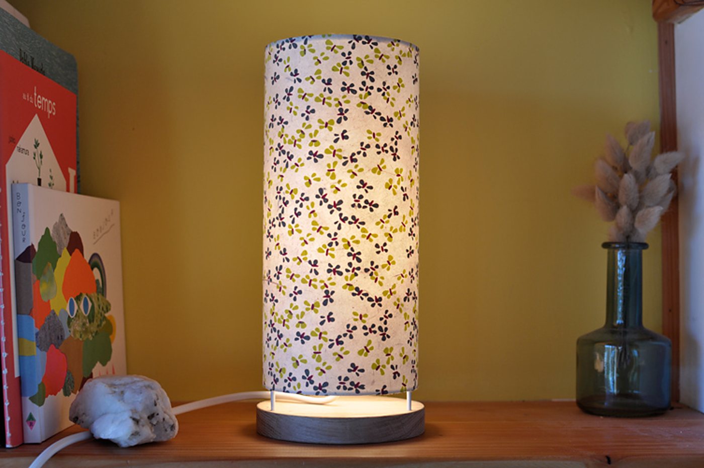 Une lampe cylindrique allumée, à l'abat-jour en tissu fleuri et au socle en bois, posée sur une étagère en bois près de livres et d'un vase en verre avec des herbes séchées.