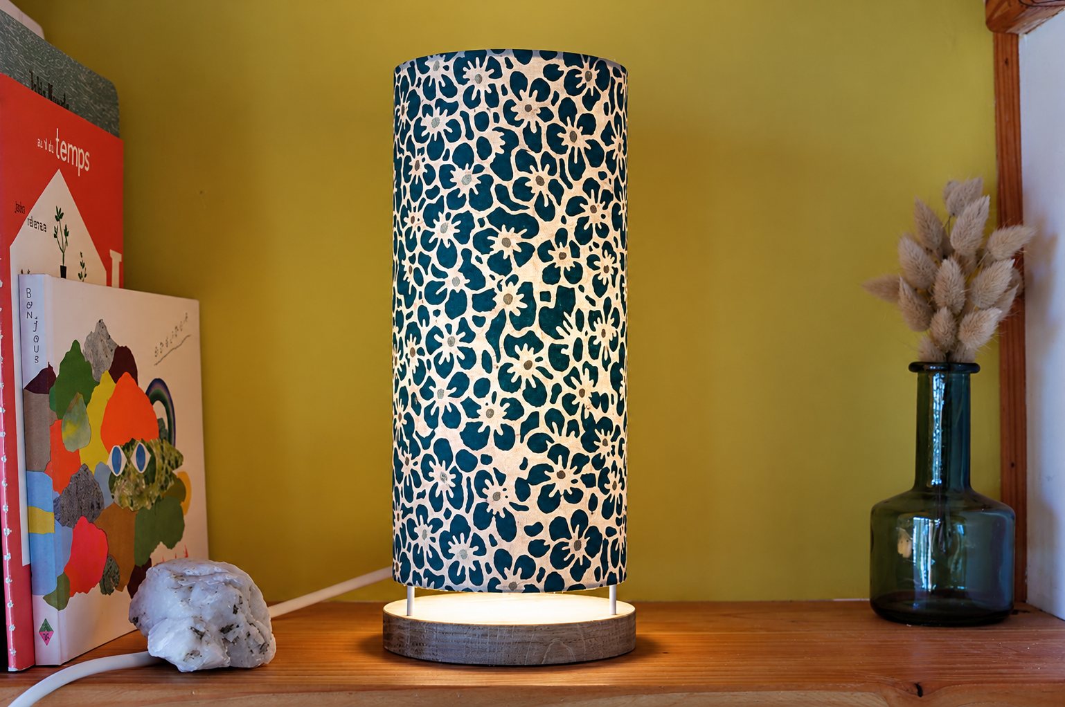 Une lampe cylindrique allumée au abat-jour à fleurs bleu canard et blanches, posée sur une étagère en bois près de livres et d'un vase en verre avec des plantes séchées.