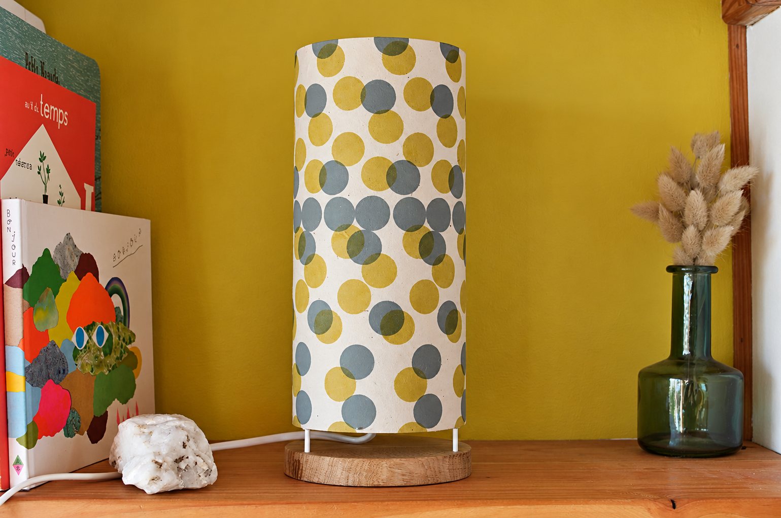 Une lampe cylindrique en papier à pois gris et jaunes, posée sur une base en bois, est placée sur une étagère entre des livres illustrés et un vase en verre avec des herbes séchées.