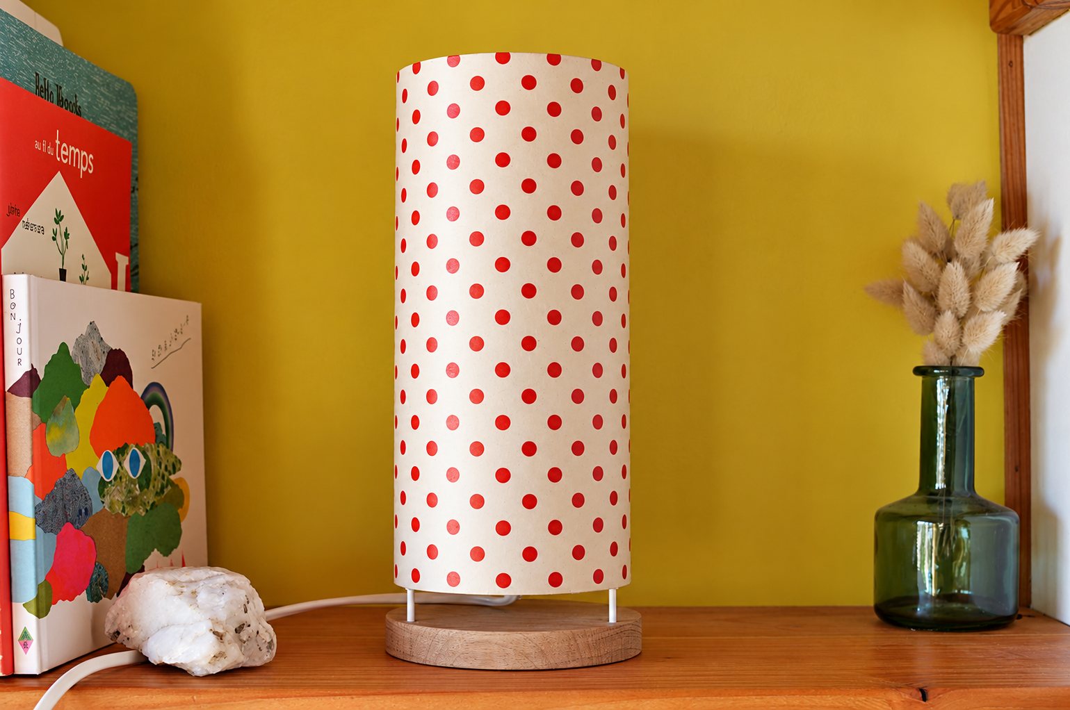 Une lampe cylindrique en papier blanc à pois rouges, sur un socle en bois clair, est posée sur une étagère devant un mur jaune, entre des livres d'enfants et un vase en verre avec des fleurs séchées.