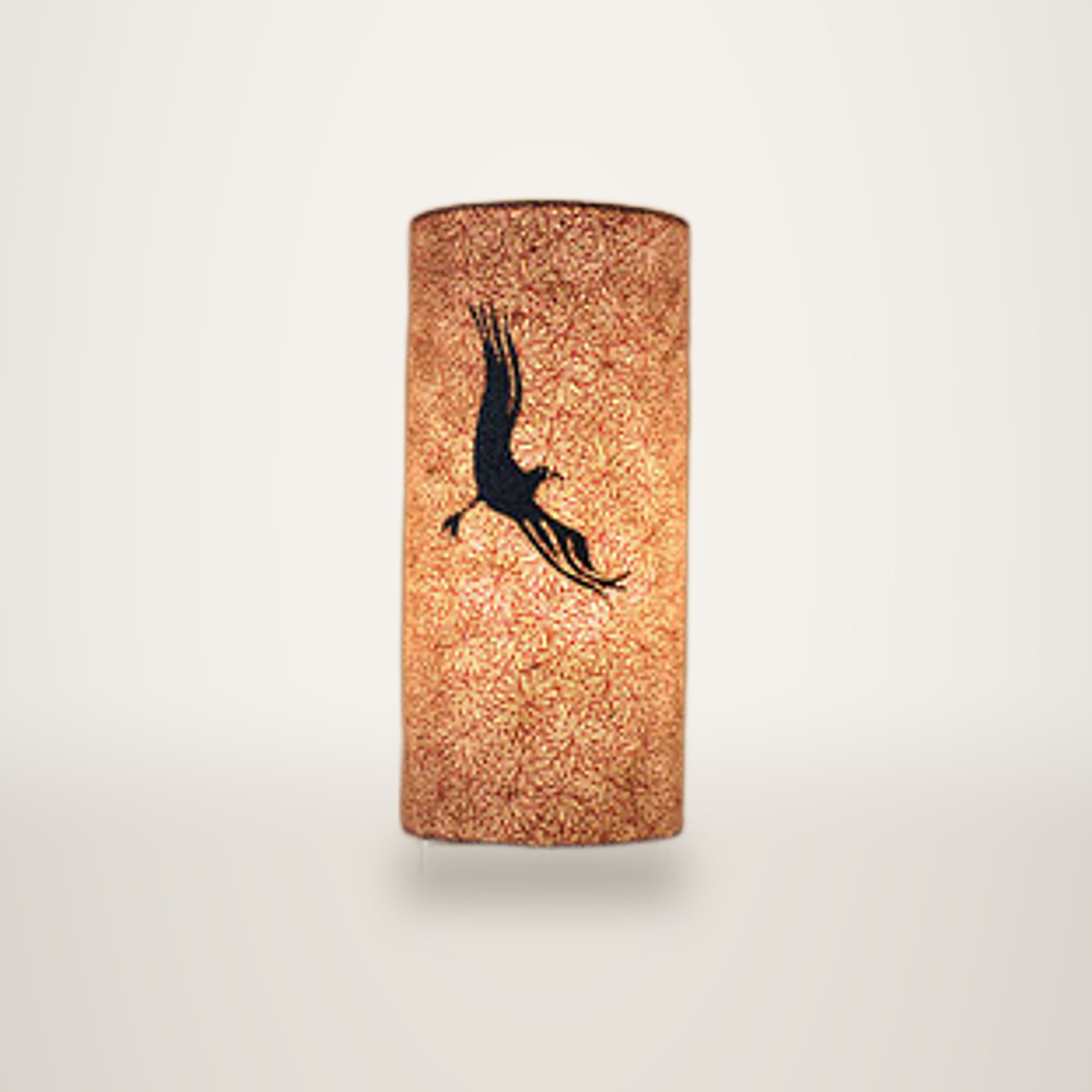 Une lampe de table cylindrique allumée, dont l'abat-jour texturé est décoré de la silhouette sombre d'un oiseau déployant ses ailes, posée sur une surface en bois à côté d'une pierre claire.